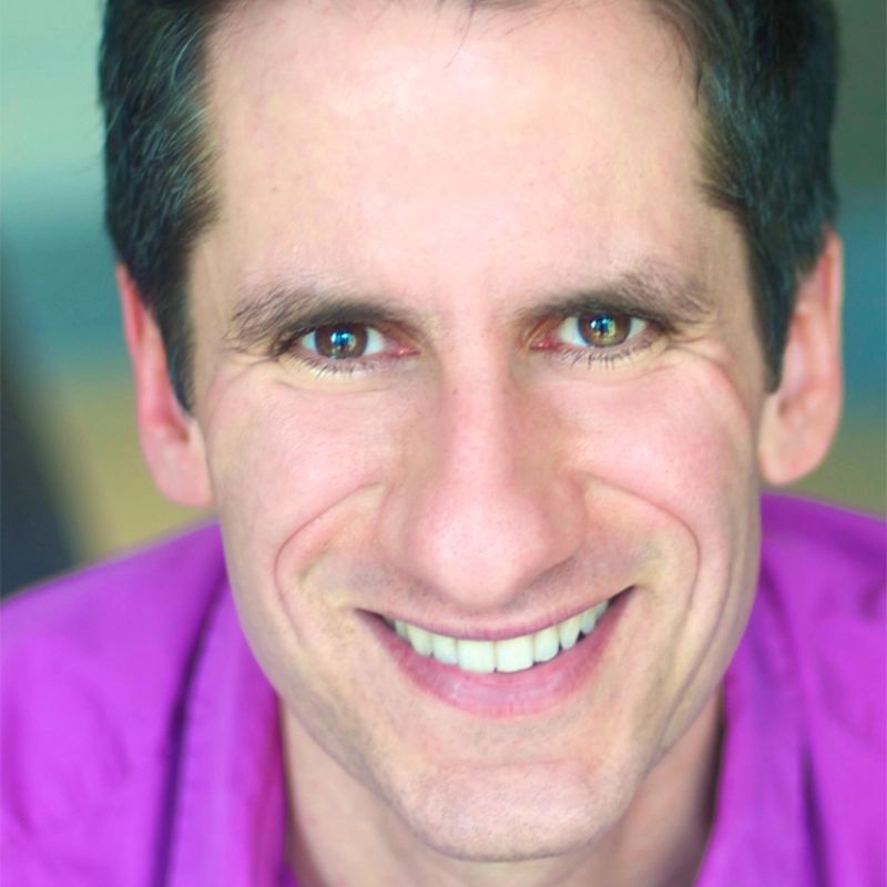 A man with short dark hair smiles broadly, showing his teeth. He has light skin and is wearing a bright purple shirt. The background is blurred, drawing focus to his happy expression and direct gaze, reminiscent of Seth Rudetsky’s vibrant energy.