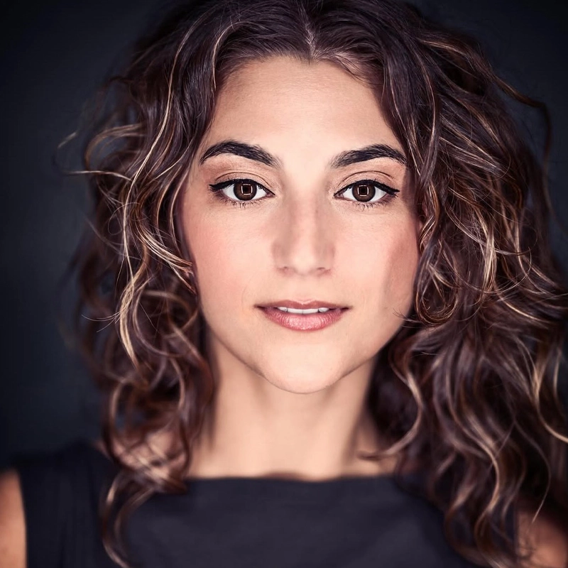 Analise Scarpaci, a woman with light skin and curly brown hair with blonde highlights, looks directly at the camera. She has brown eyes, defined eyebrows, and subtle makeup. Her dark top and the blurred background draw focus to her face.
