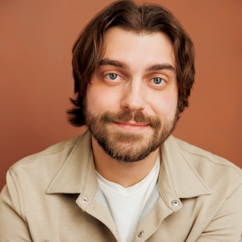 Nathan David Smith, a bearded man with medium-length brown hair and blue eyes, smiles gently at the camera. He wears a beige jacket over a white shirt. The background is a solid warm brown color.