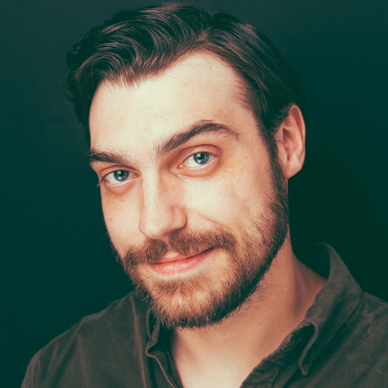 Nathan David Smith, a man with fair skin, short brown hair, and a full beard, smiles slightly at the camera. He has blue eyes and wears a dark olive green button-up shirt. The plain dark background highlights his well-lit face.