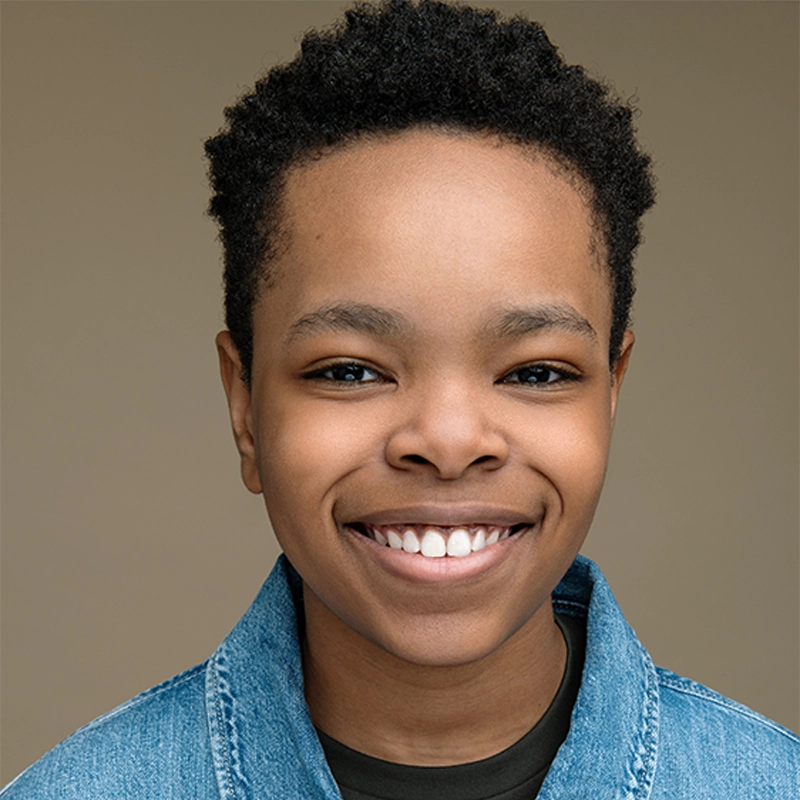 A young person with short, curly black hair and brown skin smiles brightly. They are wearing a blue denim jacket over a dark top. The plain beige background highlights their friendly expression—capturing the vibrant energy of Walter Russell III.
