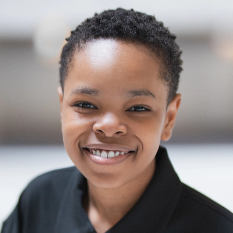 A smiling young person, Walter Russell III, with short, curly black hair wearing a black collared shirt. The softly blurred background keeps the focus on their friendly face and warm expression. The lighting is bright and even.