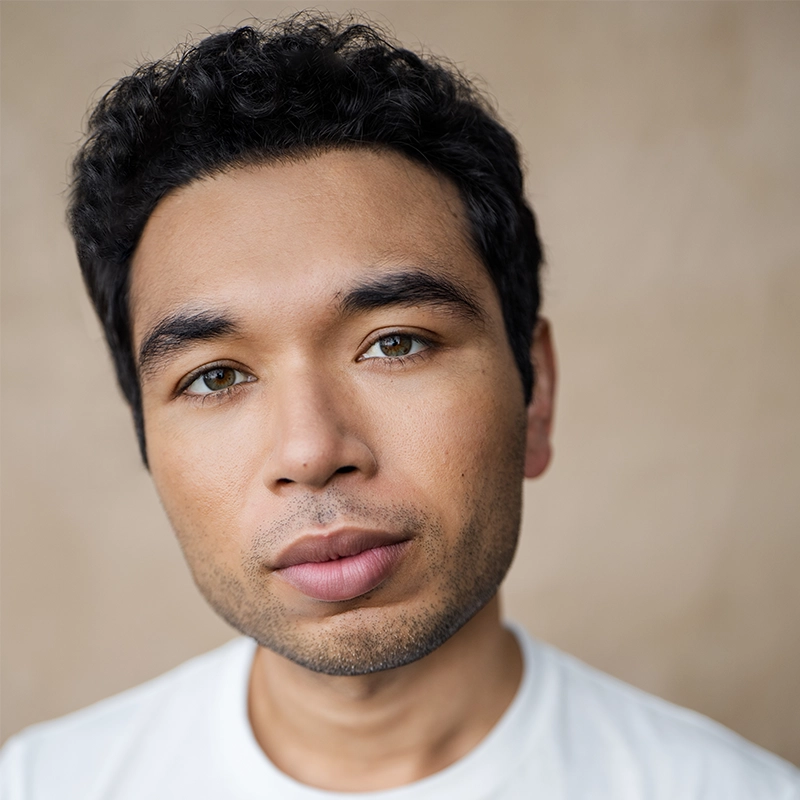 Aaron Alcaraz, a young man with short, dark curly hair and light brown skin, gazes softly at the camera. He has hazel eyes, full lips, slight stubble, and wears a white shirt against a neutral beige background.