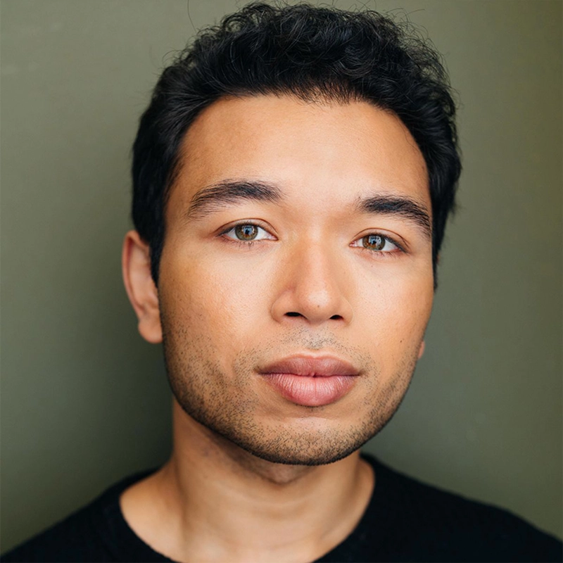 Headshot of Aaron Alcaraz, a man with short dark hair and light brown eyes, looking directly at the camera. He has a slight beard and is wearing a plain black crew-neck shirt against a solid olive-green background.