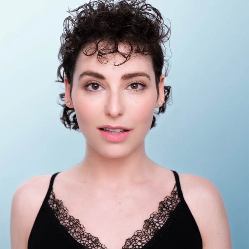 A person with short, curly, dark hair and light skin is facing the camera against a pale blue background. Ilana Gabrielle wears small earrings and a black camisole with lace trim, her expression neutral with a slight smile and natural makeup.