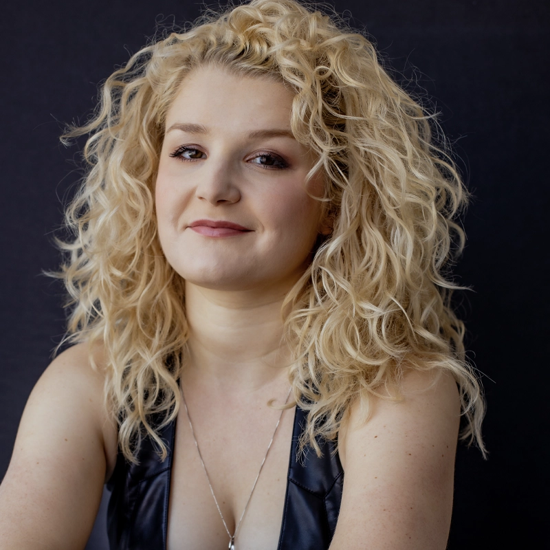 Emma Maxwell, with fair skin and voluminous curly blonde hair, smiles softly. She wears a sleeveless black leather top and a thin necklace. The solid black background highlights her hair and confident expression.