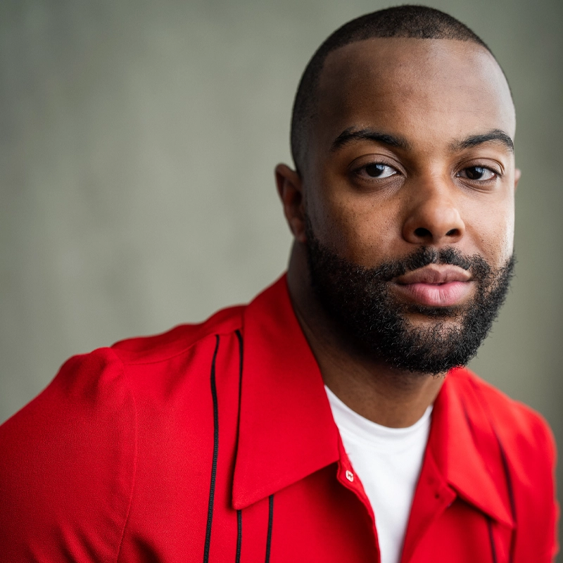 A man with a close-cropped beard and short hair wears a red collared shirt over a white t-shirt. He looks at the camera with a neutral expression. The background is blurred and gray.