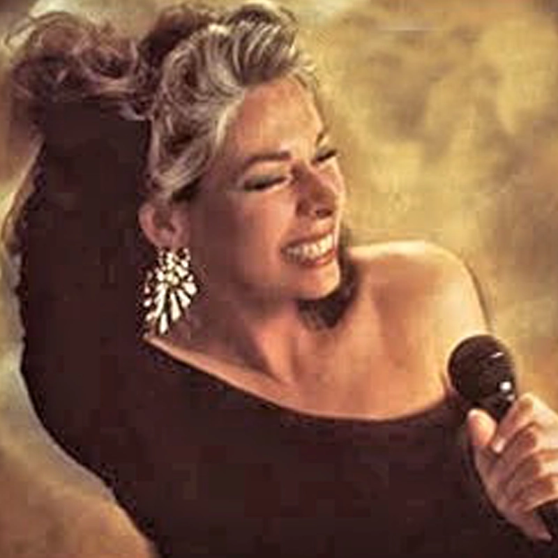 A smiling woman with gray-streaked hair and large, sparkling earrings holds a microphone. She wears a black off-the-shoulder top and has one arm raised behind her head, appearing joyful while singing against a golden, softly blurred background.