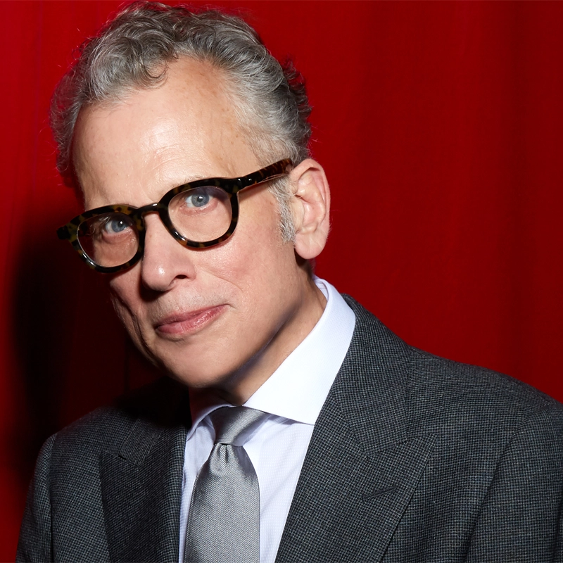 A man with short gray hair and tortoiseshell glasses wears a gray suit, white shirt, and silver tie. He stands in front of a rich red curtain, looking at the camera with a neutral expression. The lighting highlights his face and suit, capturing the polished style often associated with Billy Stritch.