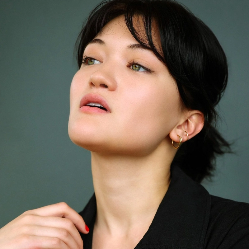 Isa Camille Briones, with short black hair and green eyes, looks slightly upward and to the side. She wears a black top, small hoop earrings, and natural makeup. Her hand rests near her collar against a plain, muted greenish-gray background.