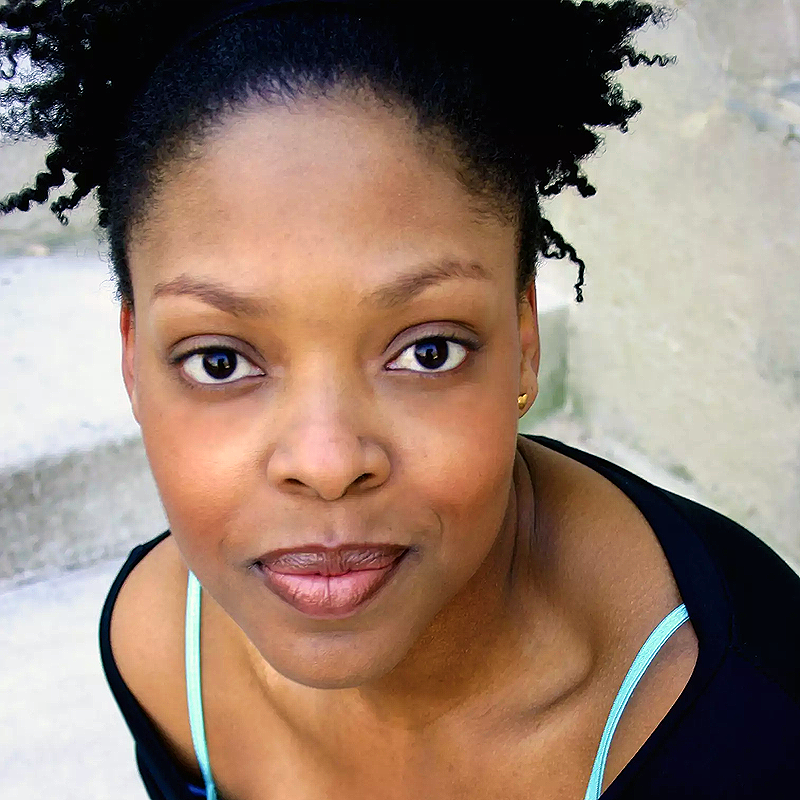 A woman with medium-dark skin and natural curly hair pulled back looks directly at the camera, slightly smiling. She wears a black top with light blue straps—Rheaume Crenshaw stands out against blurred stone steps in soft, natural light.