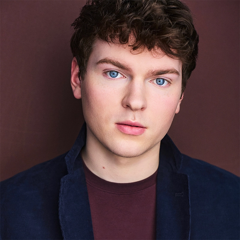 A young man with fair skin, short curly brown hair, and striking blue eyes faces the camera. Mitchell Sink wears a dark blue jacket over a maroon shirt. The solid brown background creates a simple and professional studio portrait.