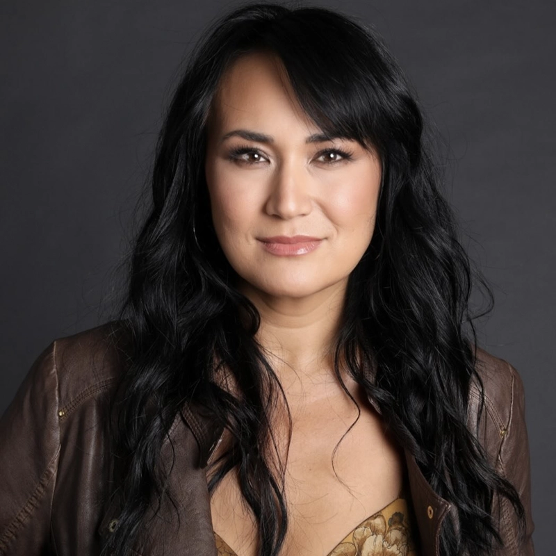 Anne Fraser Thomas smiles softly, her long, wavy black hair framing her light skin. She wears a brown leather jacket over a tan patterned top against a neutral dark gray background, with natural makeup and nude lipstick.