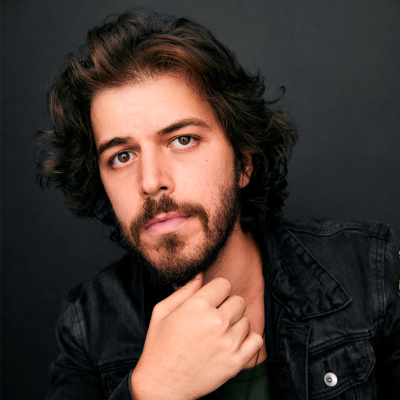 Hunter Brown, with wavy dark hair, a full beard, and mustache, poses against a dark background. He wears a black denim jacket and gazes thoughtfully at the camera, his hand resting under his chin.