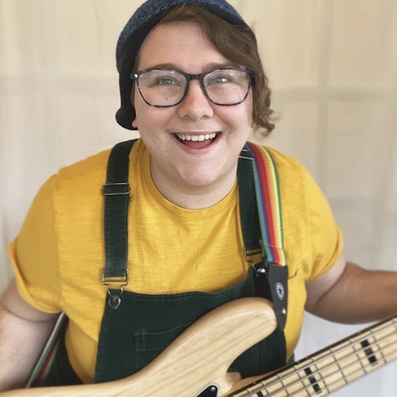 A person with short brown hair, glasses, and a beanie smiles at the camera. They wear a yellow shirt under green overalls and hold a light-colored electric bass guitar with a rainbow strap against a neutral background.