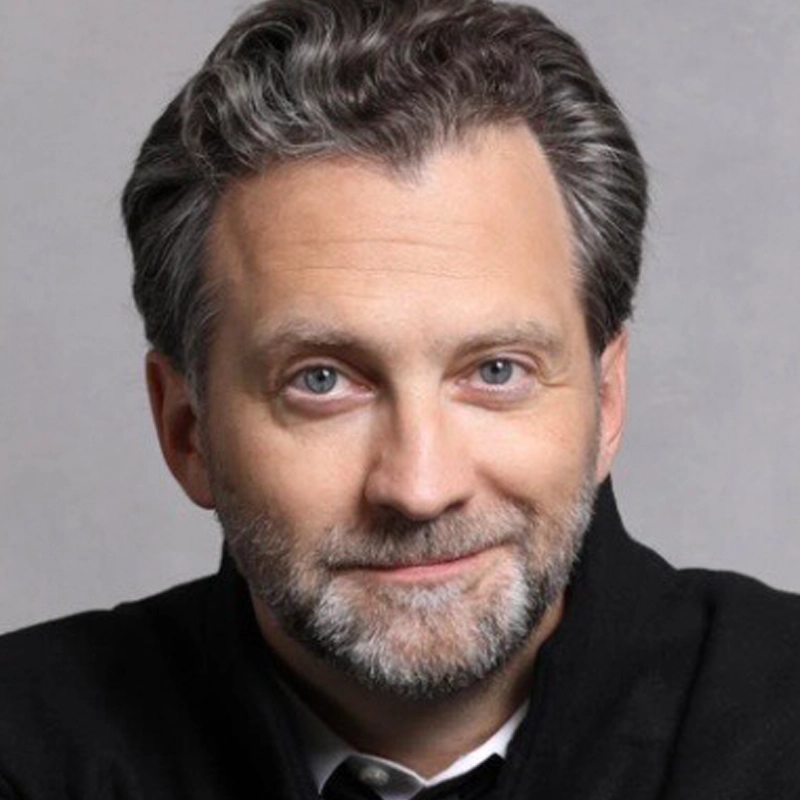A middle-aged man with light skin, blue eyes, and short, wavy salt-and-pepper hair, including a neatly trimmed beard and mustache, smiles slightly. Graham Rowat wears a dark jacket over a white shirt, posed against a plain, light gray background.