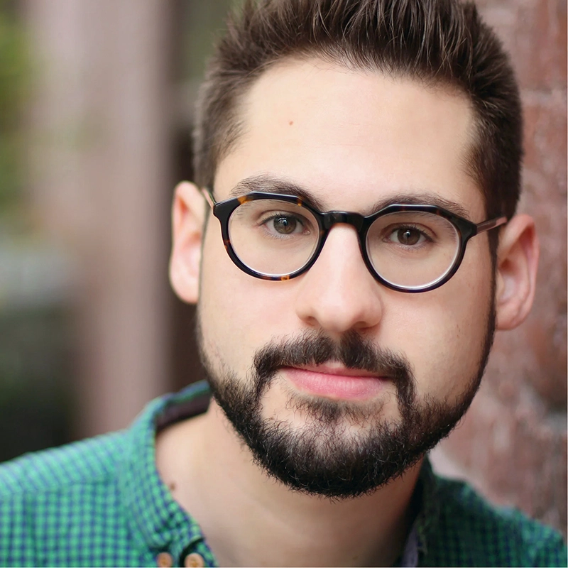 A young man with short dark hair, a trimmed beard, and round black glasses looks at the camera. He wears a green, patterned shirt and stands outdoors next to a brick wall, with a softly blurred background.