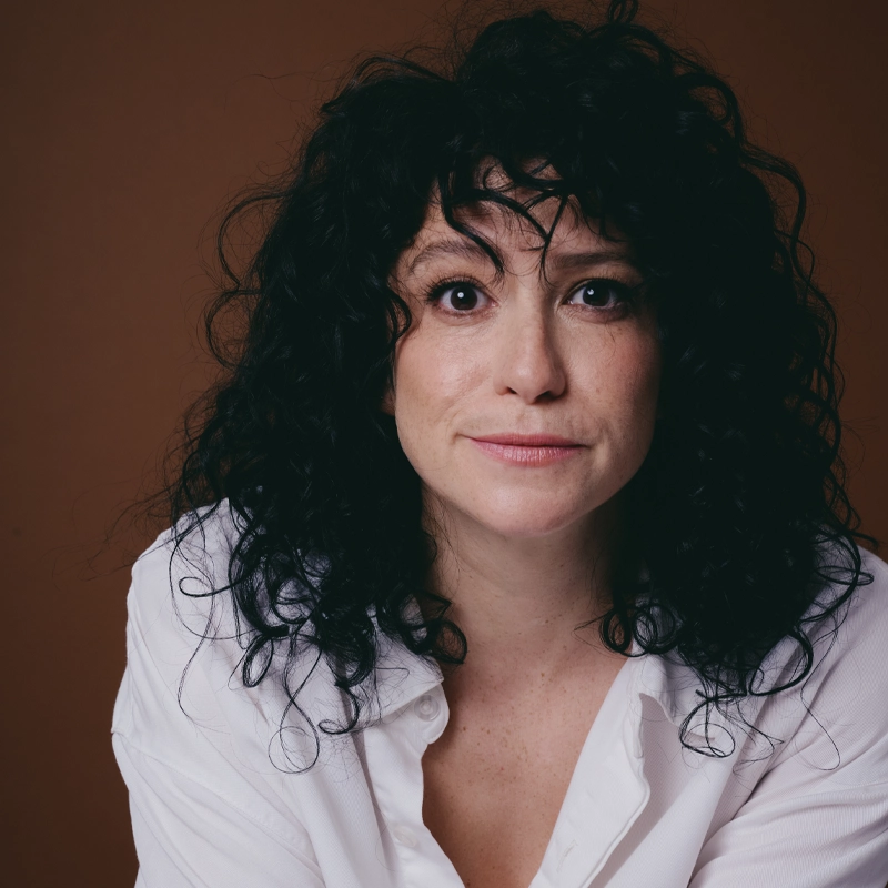 A woman with curly black hair and light skin, possibly Jordan Kai Burnett, wears a white blouse and looks directly at the camera with a neutral, slightly inquisitive expression. The plain brown background and soft lighting highlight her facial features.