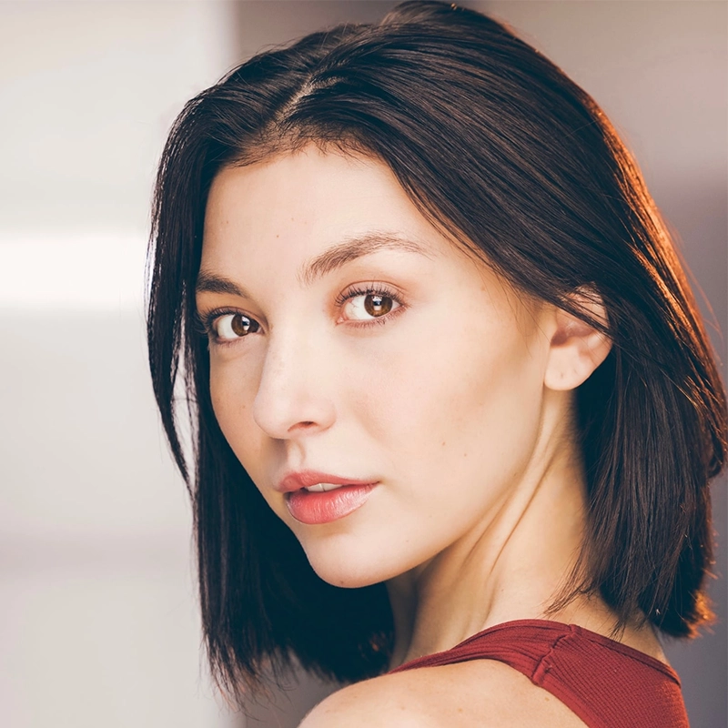 A young woman with straight, shoulder-length dark brown hair looks over her shoulder toward the camera. She has fair skin, brown eyes, and natural makeup. She wears a sleeveless reddish top and stands against a softly blurred, light-colored background.