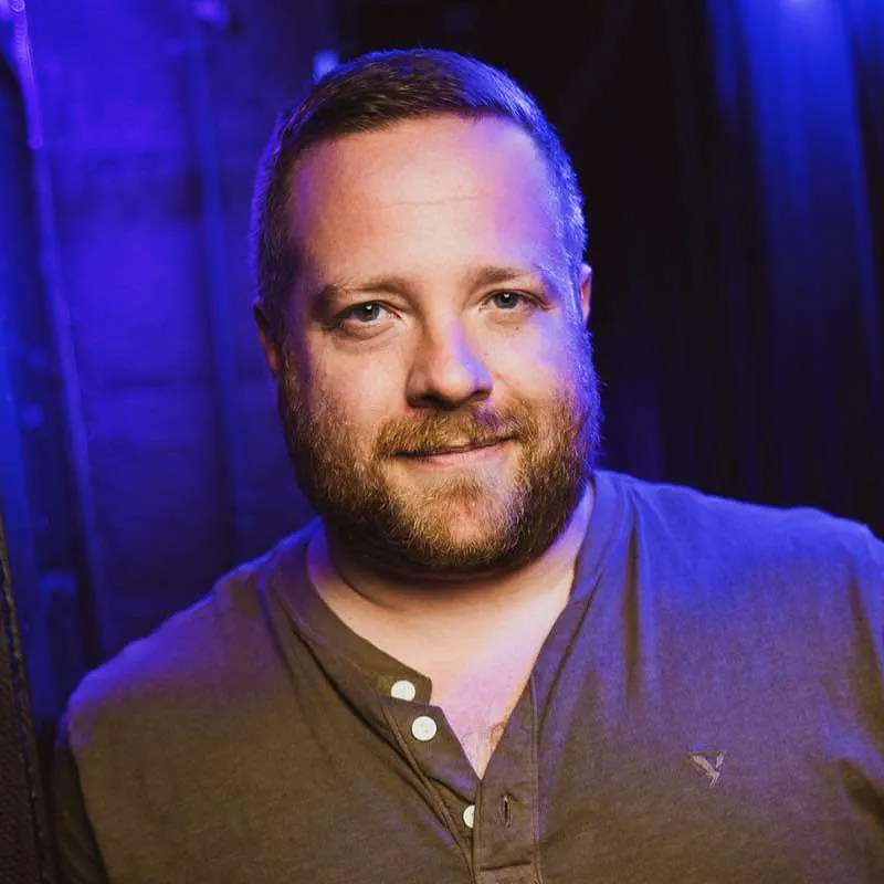 A bearded man with short brown hair stands under blue and purple lighting, wearing a buttoned brown shirt with a small bird logo. Michael Hartney smiles softly, looking directly at the camera, with a blurred dark background behind him.