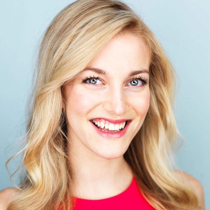 Smiling woman with long, wavy blonde hair and blue eyes wearing a bright pink sleeveless top. She is standing in front of a plain light blue background and looking directly at the camera.