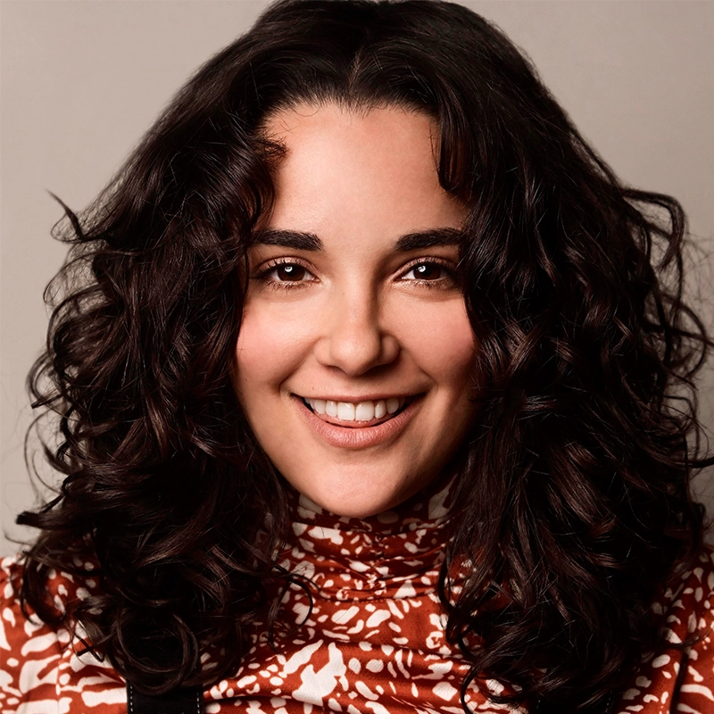 A woman with medium-length, dark curly hair smiles at the camera. She wears a high-necked, long-sleeve top with a red and white floral pattern. The background is plain and light-colored, highlighting her friendly expression.