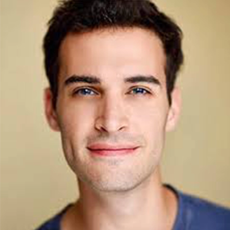 A young man with short dark hair and light skin smiles slightly at the camera. He is wearing a casual blue shirt and is posed in front of a plain light beige background. The image is well-lit, highlighting his facial features.