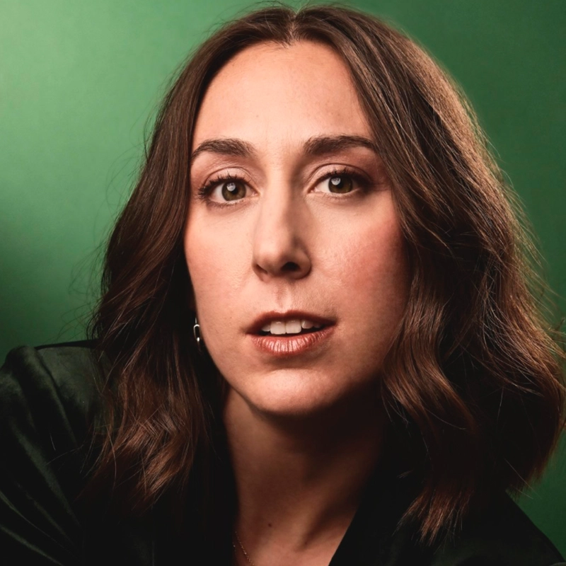 A woman with wavy brown hair and light skin looks directly at the camera against a green background. She is wearing a dark top, subtle makeup, and a neutral expression. The lighting highlights her facial features and hair.