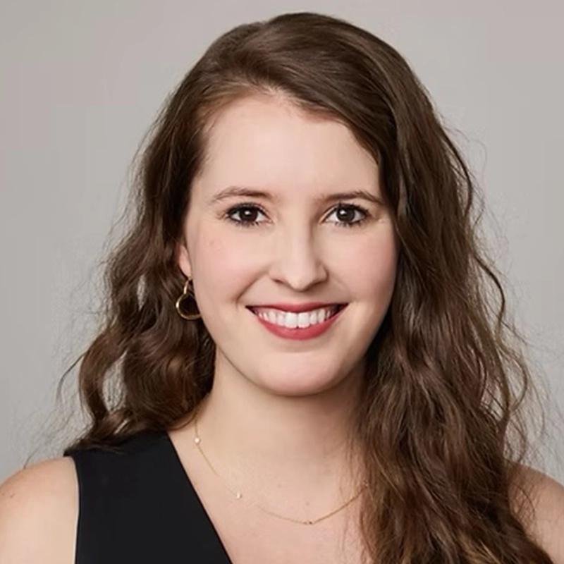 Caitlin McNeilage, a woman with long, wavy brown hair, smiles while facing the camera. She wears a black top, small hoop earrings, and a delicate necklace. The background is plain and light gray.
