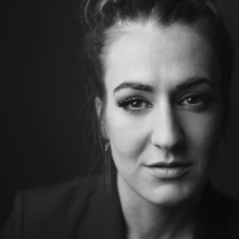 Black and white close-up portrait of Hannah Shankman, a woman with light skin, slightly smiling, looking forward. She has neatly groomed eyebrows, long eyelashes, hair pulled back, and wears a dark blazer against a plain dark background.