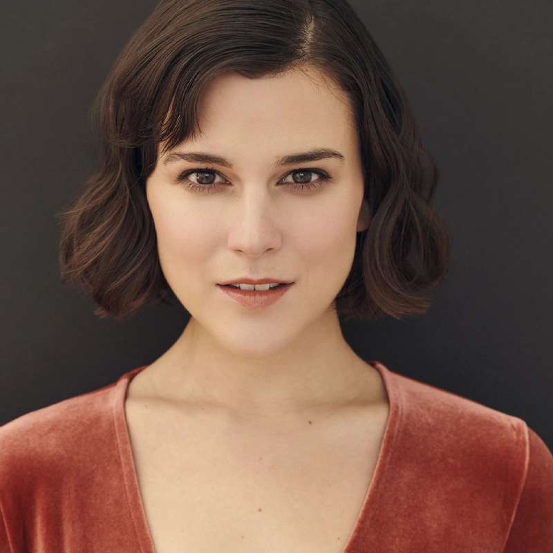 A person with short wavy brown hair and fair skin looks directly at the camera, reminiscent of Alexandra Socha, wearing a coral velvet V-neck top. The background is a plain, dark color, and the soft lighting highlights their facial features.