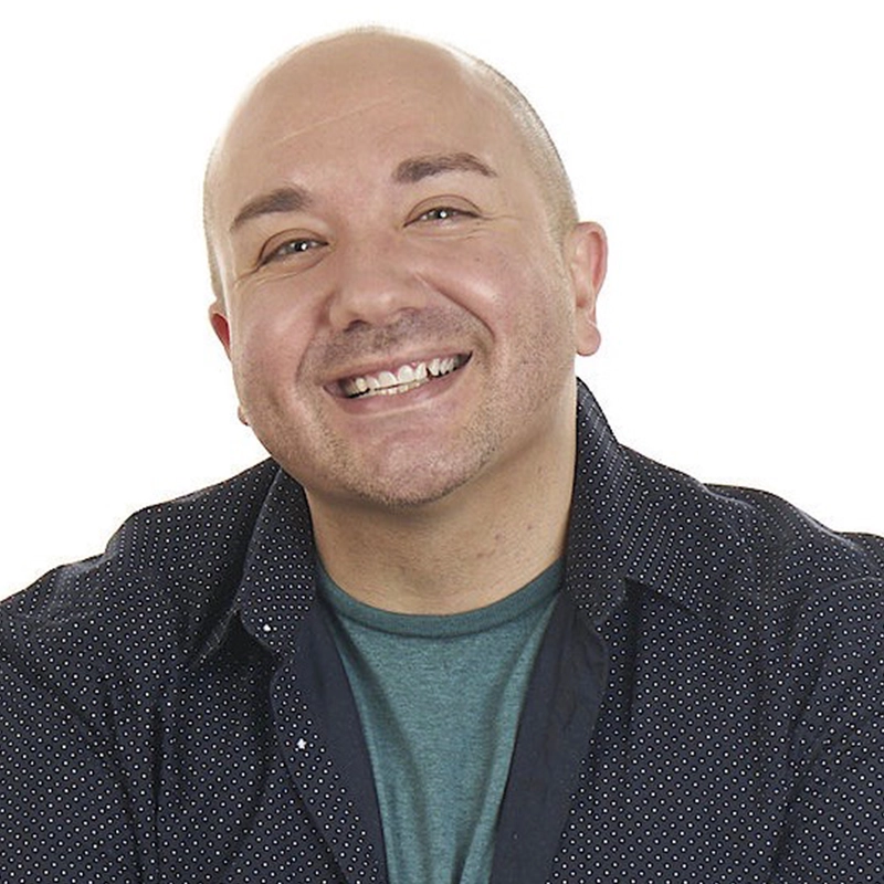 A smiling, bald person in a dark patterned button-up shirt over a teal T-shirt poses against a white background, looking directly at the camera with a friendly expression reminiscent of Janine LaManna.