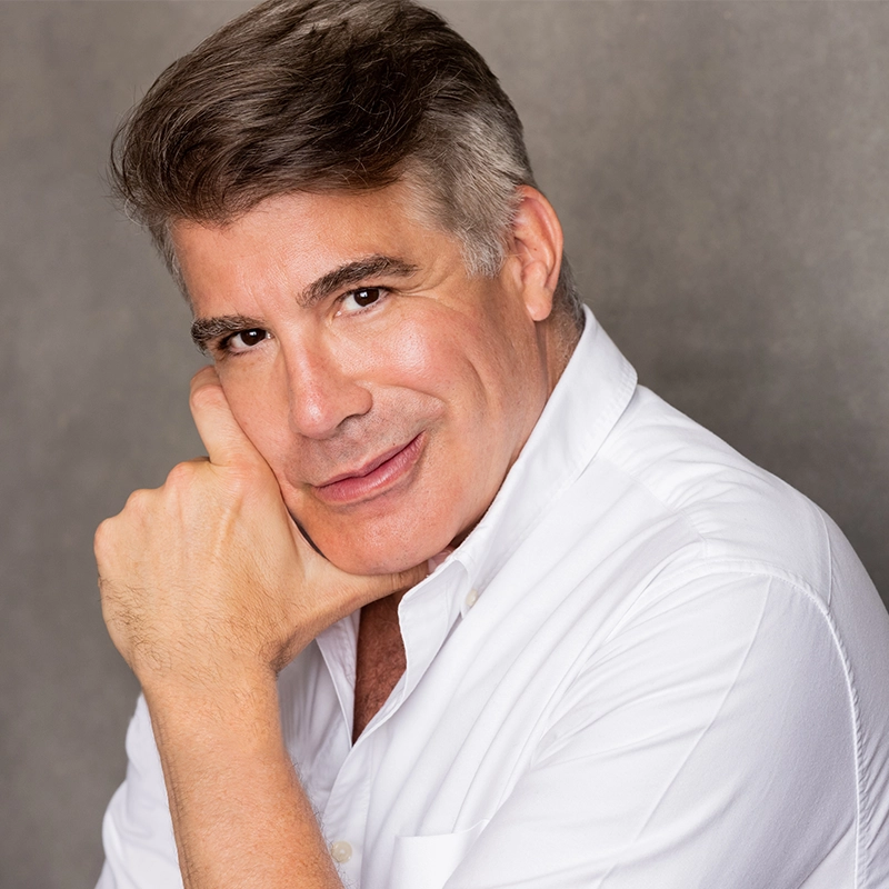 A middle-aged man with gray hair smiles gently, resting his chin on his fist. He wears a white button-down shirt and poses against a plain gray background, giving a friendly and approachable expression reminiscent of Janine LaManna’s warmth.