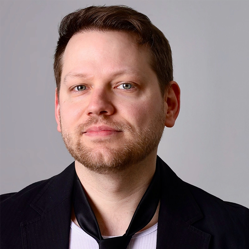 A man with fair skin, short brown hair, and a trimmed beard looks at the camera with a neutral expression. He wears a black blazer, a white shirt, and a loosely draped black tie. The background is plain light gray, evoking Monica Rosenblatt’s style.