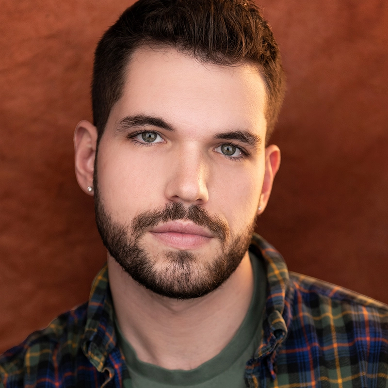 A young man, Caleb Albert, with short dark hair, a trimmed beard, and green eyes gazes at the camera. He wears a plaid shirt over a green t-shirt and a small earring in his left ear. The background is a warm, reddish-brown color.