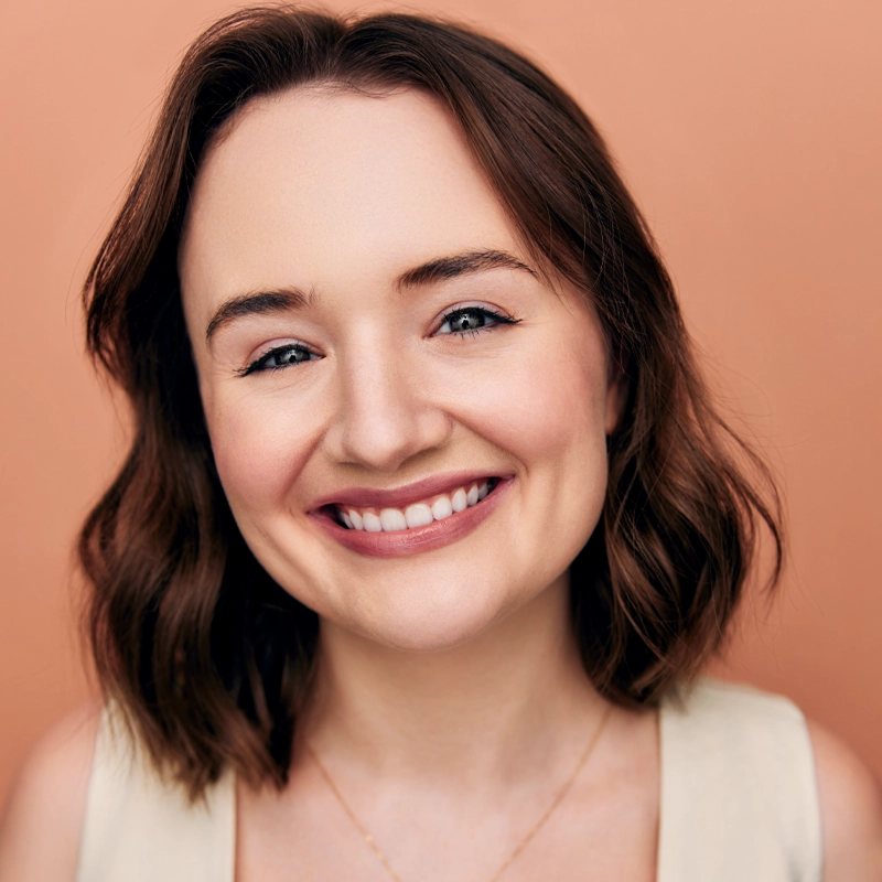 Young woman with light skin and shoulder-length brown hair smiles warmly at the camera. She wears a cream sleeveless top and a delicate gold necklace. The background is a solid, soft peach color, creating a bright and friendly portrait.