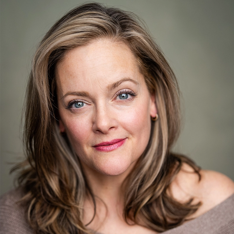 A woman with light skin and long, wavy, light brown hair smiles softly. Janine LaManna has blue eyes, subtle makeup, and wears a beige off-the-shoulder top. The blurred, neutral-toned background highlights her face.