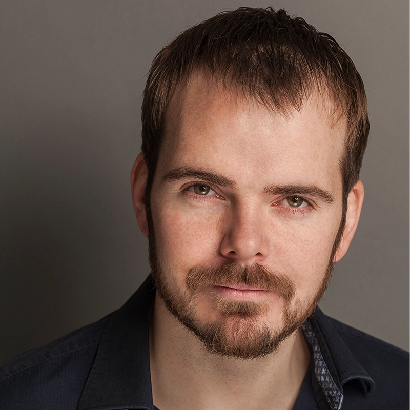 A man with short brown hair and a trimmed beard looks directly at the camera with a slight smile. He wears a dark collared shirt against a neutral gray background, softly lit to highlight his features in Monica Rosenblatt’s signature portrait style.
