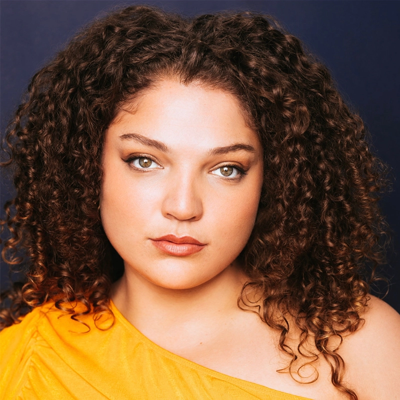 Monica Rosenblatt, with fair skin and voluminous curly brown hair, looks directly at the camera. She wears a one-shoulder yellow top and natural makeup, posing against a dark blue background with a calm, confident expression.