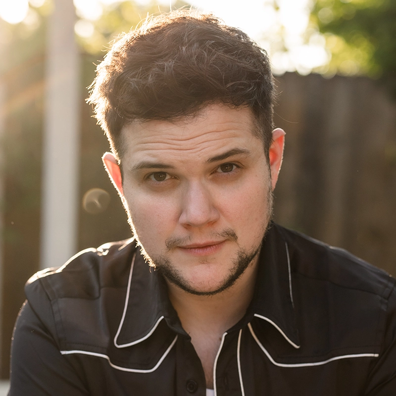A young man with short, wavy dark hair and light skin gazes confidently at the camera. He has some facial hair and wears a black collared shirt with white piping. Sunlight softly illuminates Preston Max Allen, with a blurred wooden fence in the background.