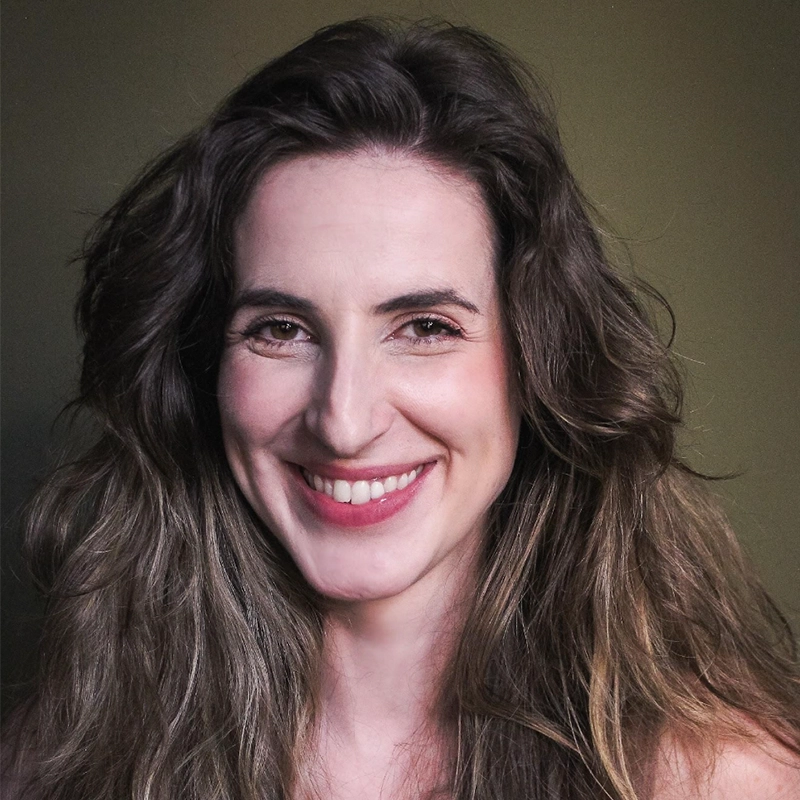 Monica Rosenblatt, a woman with long, wavy brown hair and fair skin, smiles warmly at the camera. She wears light makeup with pink lipstick and stands against a plain, muted green background, her expression friendly and approachable.
