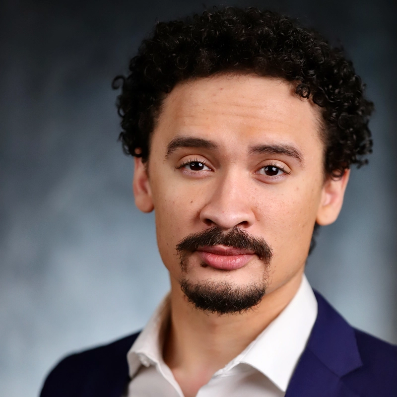 A man with short curly hair and a goatee, reminiscent of Miles Davis, wears a dark blue suit jacket and white shirt. He looks confidently at the camera, the blurred background drawing focus to his expressive face and professional attire.