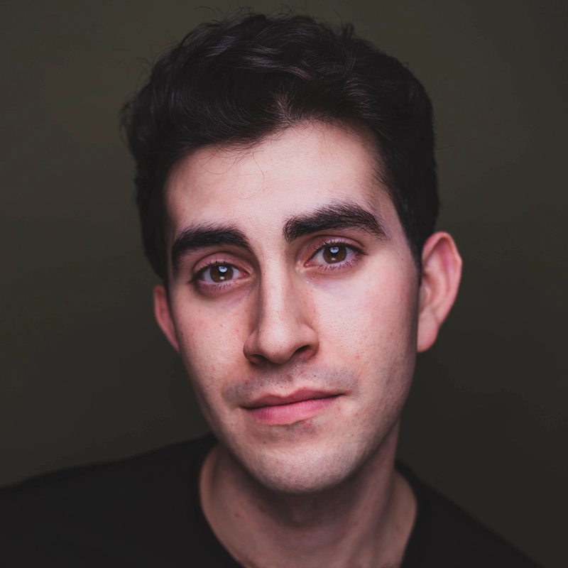 A young man with short, dark hair and thick eyebrows gazes softly at the camera. He has fair skin, dark brown eyes, and a hint of a smile. The background is plain dark green, and Justin Albinder wears a black shirt under soft, even lighting.
