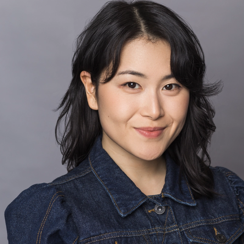 A woman with medium-length wavy black hair smiles softly at the camera. She is wearing a dark denim jacket with a buttoned collar. The background is a plain, light gray, giving a professional and friendly headshot appearance.