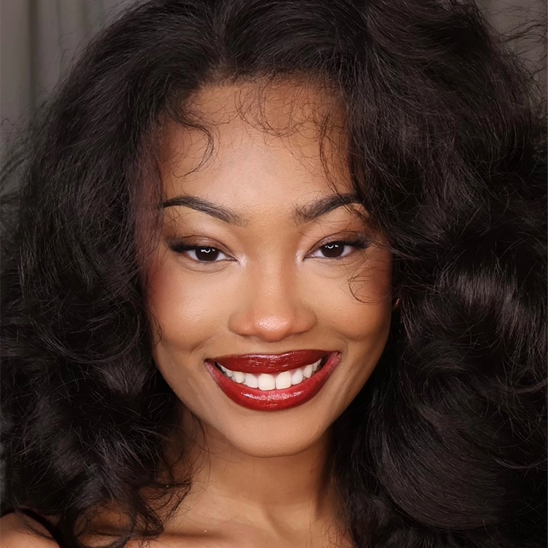 A woman with voluminous, wavy dark hair smiles confidently. She has smooth skin, bold red lipstick, neatly shaped eyebrows, and subtle eye makeup. The background is softly blurred, keeping the focus on her radiant face.