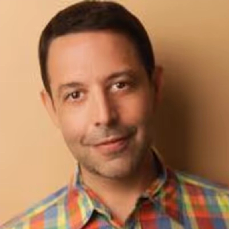 Justin Albinder, with short dark hair and light skin, is smiling softly in a colorful plaid shirt. The plain warm beige background adds to the friendly, approachable feel, while even lighting highlights Justin’s face.