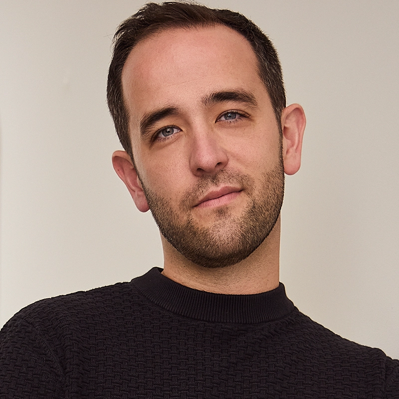 A man with short brown hair, trimmed beard, and fair skin wears a textured black sweater. He stands against an off-white background, looking at the camera with a relaxed, confident expression and a slight smile.