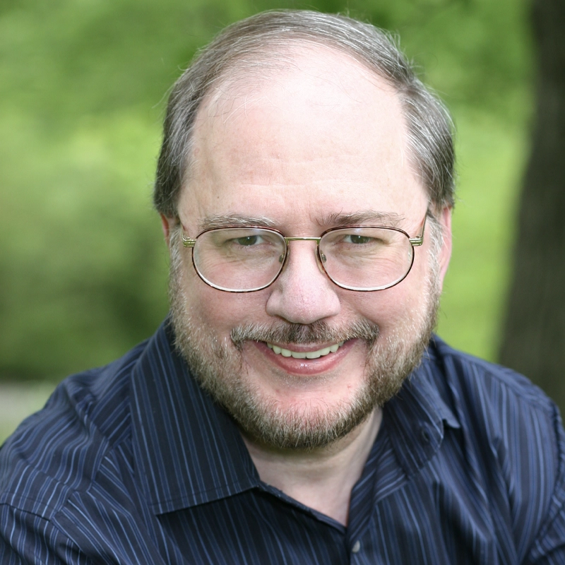 A middle-aged man with light skin, short graying hair, glasses, and a trimmed beard smiles at the camera. Reminiscent of Rupert Holmes, he wears a dark blue pinstriped shirt against a blurred green background with trees.