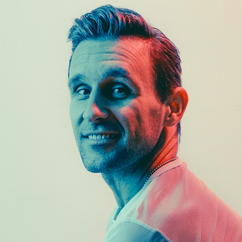 A man with short, slicked-back hair and light stubble smiles slightly while looking over his shoulder. The image of Nicolas King features a blue and pink color filter, and he wears a light shirt with a thin necklace against a plain background.