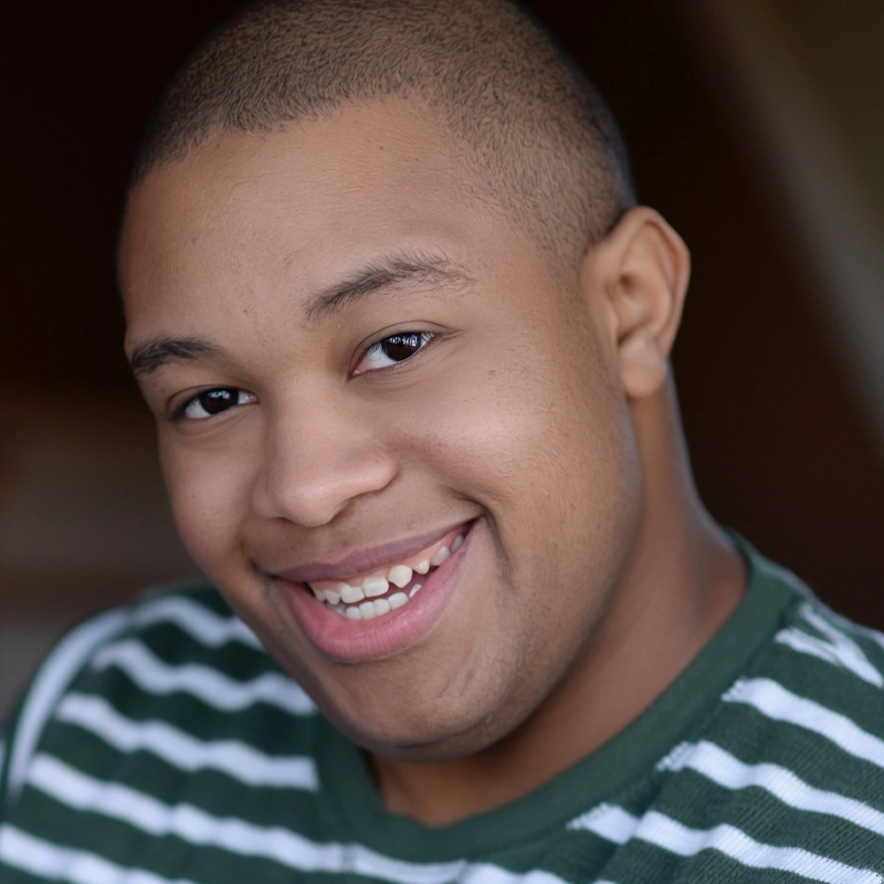 Terrence Berry, a young man with short hair, smiles warmly at the camera. He is wearing a green and white striped shirt. The background is softly blurred, keeping focus on his friendly expression.